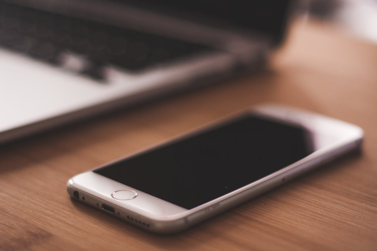 A smartphone lies on a wooden table next to a blurred laptop in a cozy indoor setting.